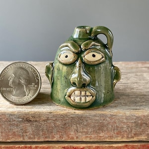 May include: A green ceramic jug with a face sculpted on the front. The jug has a handle and a spout. The face has large eyes, a prominent nose, and a wide mouth with teeth. The jug is on a wooden surface.