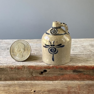 May include: A small, tan ceramic jug with a dark blue dragonfly design. The jug has a handle and a small opening at the top. A U.S. quarter is next to the jug for size comparison. The jug is sitting on a wooden surface.