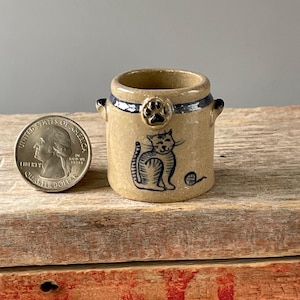 May include: A small, tan ceramic container with a blue cat illustration and a paw print. The container has a dark blue band around the top and two small handles. A quarter is next to the container for size reference.