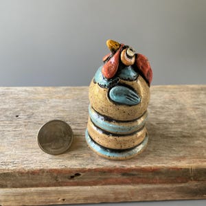 May include: A small, ceramic rooster figurine with a speckled tan body, blue accents, and red comb. The rooster is perched on a wooden surface next to a quarter coin. The figurine has a unique, handmade appearance.