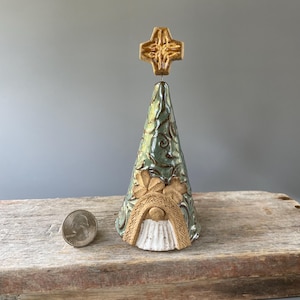 May include: A ceramic gnome Christmas tree with a cross-shaped topper. The tree is cone-shaped with a green and brown glaze, featuring leaf and swirl designs. The gnome has a white beard and a brown hat. A quarter is next to the tree.