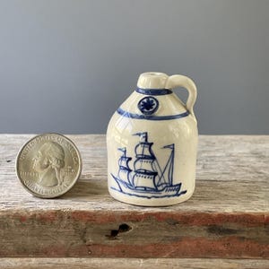 May include: Small, off-white ceramic jug with a blue ship illustration. The jug has a handle and a blue floral design near the top. A U.S. quarter is next to the jug for size comparison. The jug is on a wooden surface.