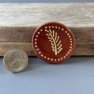 May include: A round, reddish-brown ceramic button with a yellow leaf design in the center. The button is approximately 1 inch in diameter and has a dotted border. A US quarter is next to the button for size comparison.