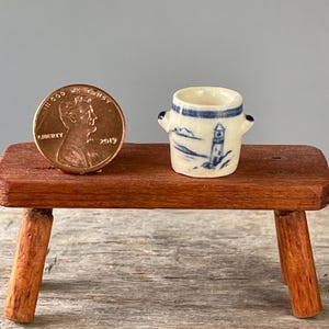 May include: A miniature ceramic pot with two small handles, featuring a blue hand-painted lighthouse design on a cream-colored background. The pot is positioned next to a standard United States copper penny for scale, showing its small size.