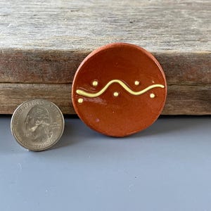 May include: A small, round, terracotta-colored ceramic piece with a yellow wavy line and dots design. A U.S. quarter is next to the ceramic piece for size comparison. The ceramic piece is on a gray surface.