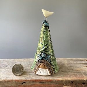 May include: A ceramic sculpture of a cone-shaped figure with a bird on top. The cone is green with blue floral accents and a textured pattern. The bird is off-white. A quarter is next to the sculpture.
