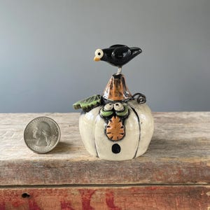 May include: A whimsical ceramic Halloween pumpkin figurine with a black bird perched on top. The pumpkin is off-white with a carved face, green leaf, and orange accents. A US quarter is next to the figurine for size reference.
