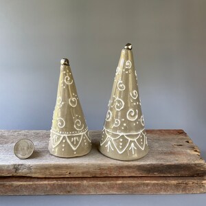 May include: Two ceramic cone-shaped Christmas trees with white painted designs and silver bells on top. The trees are on a wooden surface.