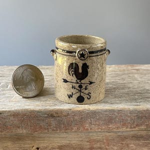 May include: A small, tan ceramic jar with a rooster and compass rose design. The jar has a black rim and a star accent. The jar is approximately 3 inches tall and 2.5 inches wide.
