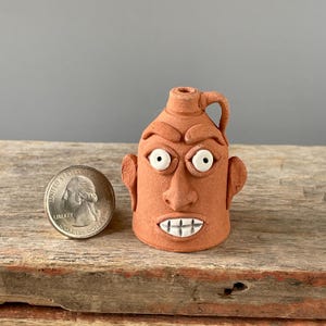 May include: A small, terracotta-colored ceramic jug with a face sculpted on the front. The face has white eyes, a nose, ears, and a mouth with white teeth. A U.S. quarter is next to the jug for size comparison. The jug sits on a weathered wooden surface.