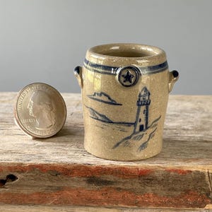 May include: A miniature tan stoneware crock with two small side handles, featuring a blue painted lighthouse scene and a star emblem. A United States quarter coin is placed next to the crock to provide a clear sense of its small scale.