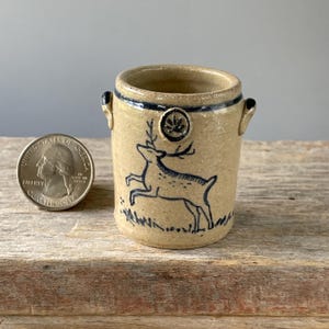 May include: A small, tan ceramic jar with a dark blue deer illustration. The jar has a dark blue rim and handles. A quarter is next to the jar for size reference. The jar is on a wooden surface.