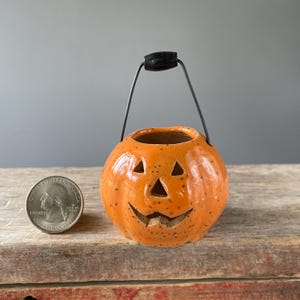 May include: A small, orange ceramic pumpkin bucket with a carved face, black handle, and a black handle. The pumpkin has a speckled texture and is sitting on a wooden surface. A quarter is next to the pumpkin.