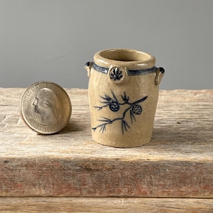 May include: Small, tan ceramic jar with a blue floral design and two small handles. The jar has a dark blue rim and a decorative blue floral accent. A U.S. quarter is next to the jar for size comparison.