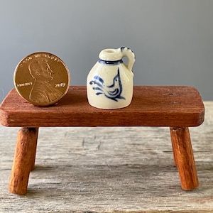 May include: Miniature ceramic jug with a blue bird design and handle. The jug is cream-colored with blue accents and is next to a penny for scale. The jug is on a small wooden table.