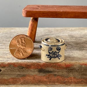 May include: A miniature beige ceramic crock with a lid, featuring a blue hand-painted crab design and two small side handles. The item is positioned next to a copper-colored United States one-cent coin for scale, showing its small size.