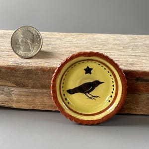 May include: A small, round, yellow ceramic dish with a scalloped, rust-colored rim. The center features a black bird silhouette and a small black star, surrounded by a dotted circle. A U.S. quarter coin is placed nearby for scale reference.