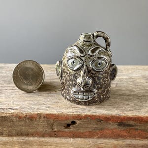 May include: A small, handcrafted ceramic jug with a face sculpted on the front. The jug is brown with speckled detailing, has a handle, and is next to a quarter. The face has large eyes, a nose, and a smiling mouth.