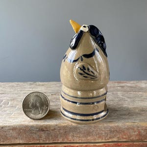 May include: A ceramic rooster figurine with a tan body, blue accents, and a yellow beak. The rooster has blue stripes around the base and decorative blue wing details. A US quarter is next to the figurine for size comparison. The figurine is on a wooden surface.