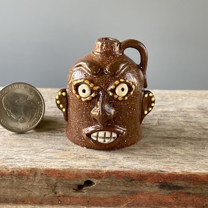 May include: A brown ceramic jug with a face sculpted on the front. The jug has a handle and a spout. The face has large eyes with yellow accents, a nose, and a mouth with teeth. A quarter is next to the jug.
