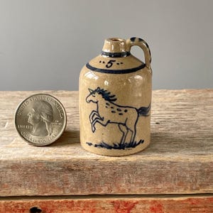 May include: A small, tan ceramic jug with a blue horse illustration and the number "5". The jug has a handle and a dark blue band around the top. A U.S. quarter is next to the jug for size comparison.