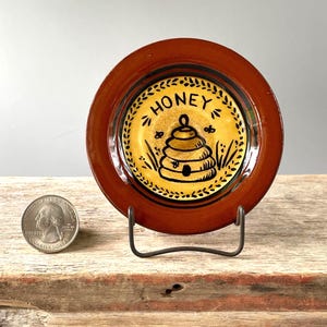 May include: A small, round, brown ceramic plate with a yellow center featuring a black and white illustration of a beehive and the word "HONEY". The plate is displayed on a metal stand. A U.S. quarter is next to the plate.