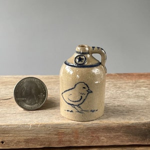 May include: A small, brown ceramic jug with a blue star and a blue and white chick design. The jug has a handle and is about the size of a US quarter.