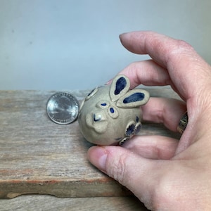May include: A small ceramic bunny figurine with blue and white painted details. The bunny has a round body and large ears. The figurine is about the size of a quarter.