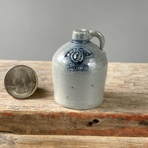 May include: A miniature light gray ceramic jug with a single handle and a glossy finish. The front features a stamped circular emblem with a star and the text Tiny Wren. A United States quarter coin is placed next to the jug for scale.