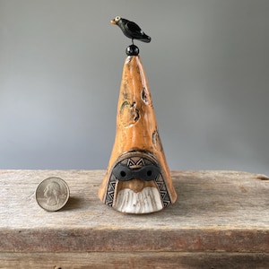 May include: A handcrafted ceramic sculpture featuring a conical form with a textured, orange-brown surface. A black bird figurine sits atop a black sphere at the sculpture's apex. The base has a face-like design with a white beard and black eye mask.