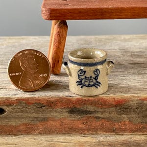 May include: A miniature beige ceramic crock featuring a blue hand-painted crab design and a blue band around the rim, positioned next to a copper-colored United States one-cent coin for scale, showing the small size of the vessel.