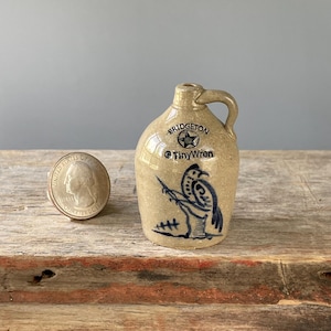 May include: A small tan ceramic jug with a handle, featuring a blue hand-painted bird illustration and the text "BRIDGETON" with a star and "TinyWren". A United States quarter coin is placed next to the jug for a clear sense of scale.