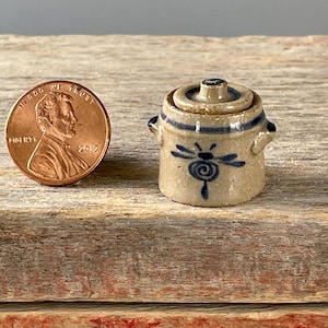 May include: A miniature tan ceramic pot with a lid, adorned with blue dragonfly designs and stripes. The pot has small handles and is next to a penny for size comparison. The pot is on a wooden surface.