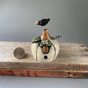 May include: A ceramic figurine shaped like a white pumpkin with a brown stem, a green leaf, and a black bird perched on top. The pumpkin features a face with green eyes, an orange nose, and a black dot mouth. A US quarter coin is placed beside it.