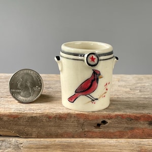 May include: A small, cream-colored ceramic vessel with two side handles, featuring a hand-painted red cardinal bird and a small red star emblem near the rim. A United States quarter coin is placed next to the vessel for scale comparison.
