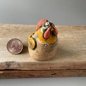 May include: A small ceramic chicken figurine with a yellow, brown, and white glaze. The chicken has a red comb and wattle, and its eyes are painted green. The figurine is about the size of a quarter.