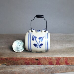 May include: A small white ceramic barrel with a blue and white floral design and a wire handle with a black plastic top. The barrel is sitting on a wooden surface.