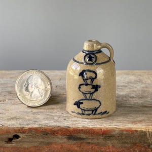 May include: A miniature tan stoneware jug with a single handle and a narrow neck. The jug features a dark blue hand-painted design of three stacked sheep and a small star near the top. A US quarter is placed next to the jug for scale reference.