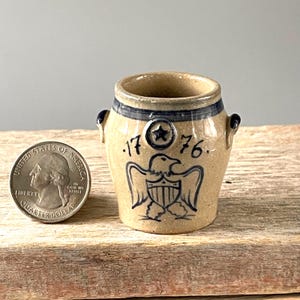 May include: Small, tan ceramic crock with blue accents and handles. Features a blue star and the year "1776" with an eagle emblem. A U.S. quarter is next to the crock for size comparison.
