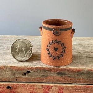 May include: Small, terracotta-colored ceramic container with a black wreath and heart design. The container has a dark rim and two small handles. A quarter is next to the container for size reference.