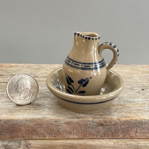 May include: A miniature tan ceramic pitcher with a blue floral design and a matching bowl. The pitcher has a handle and is decorated with blue stripes. A U.S. quarter is next to the pitcher and bowl.