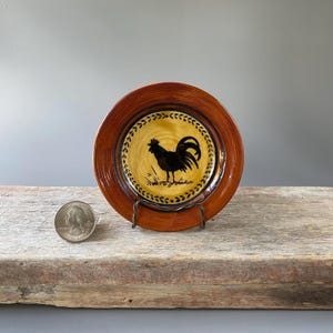 May include: A small, round decorative plate with a rooster design. The plate has a brown rim and a yellow center with a black rooster silhouette. The plate is approximately 5 inches in diameter.