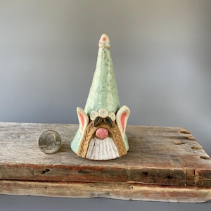May include: A ceramic gnome figurine with a green and white speckled hat, pink ears, and a pink nose. The gnome is wearing a white flower crown and has brown hair.