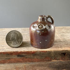 May include: A miniature brown ceramic jug with a single handle and a small circular emblem featuring a star on the front. The jug is positioned next to a United States quarter coin for scale, showing the small size of the pottery piece.
