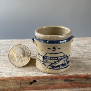 May include: A small tan stoneware crock with two side handles and a blue painted design featuring a boat, animals, and the number 5. A United States quarter coin is placed next to the crock to provide a clear sense of its small scale.