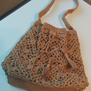 May include: A brown crochet shoulder bag with a floral pattern. The bag has a drawstring closure, a long strap, and decorative tassels with wooden beads. The bag is made of a woven material.