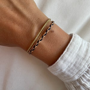 May include: A set of three bracelets on a wrist. The bracelets are made of braided string in black, beige, and gold. The gold bracelet is a delicate chain.