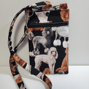 May include: Black crossbody bag with a poodle pattern. The bag features a zipper closure and a patterned strap. The poodle print includes various colors of poodles on a black background. The strap has a black, white, and brown pattern.