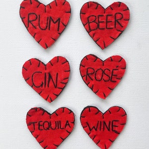 May include: Eight red fabric heart-shaped labels with black stitching and drink names: Vodka, Cider, Rum, Beer, Gin, Rosé, Tequila, and Wine. The background is white.