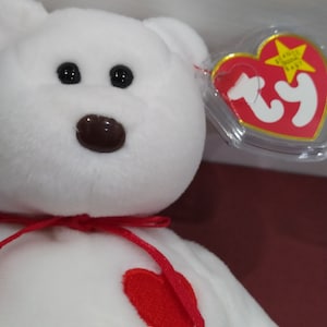 Puede incluir: Oso de peluche blanco con un corazón rojo en el pecho y una cinta roja alrededor del cuello. El oso tiene una nariz marrón y ojos negros. El oso lleva una etiqueta Ty con un corazón rojo y una estrella.